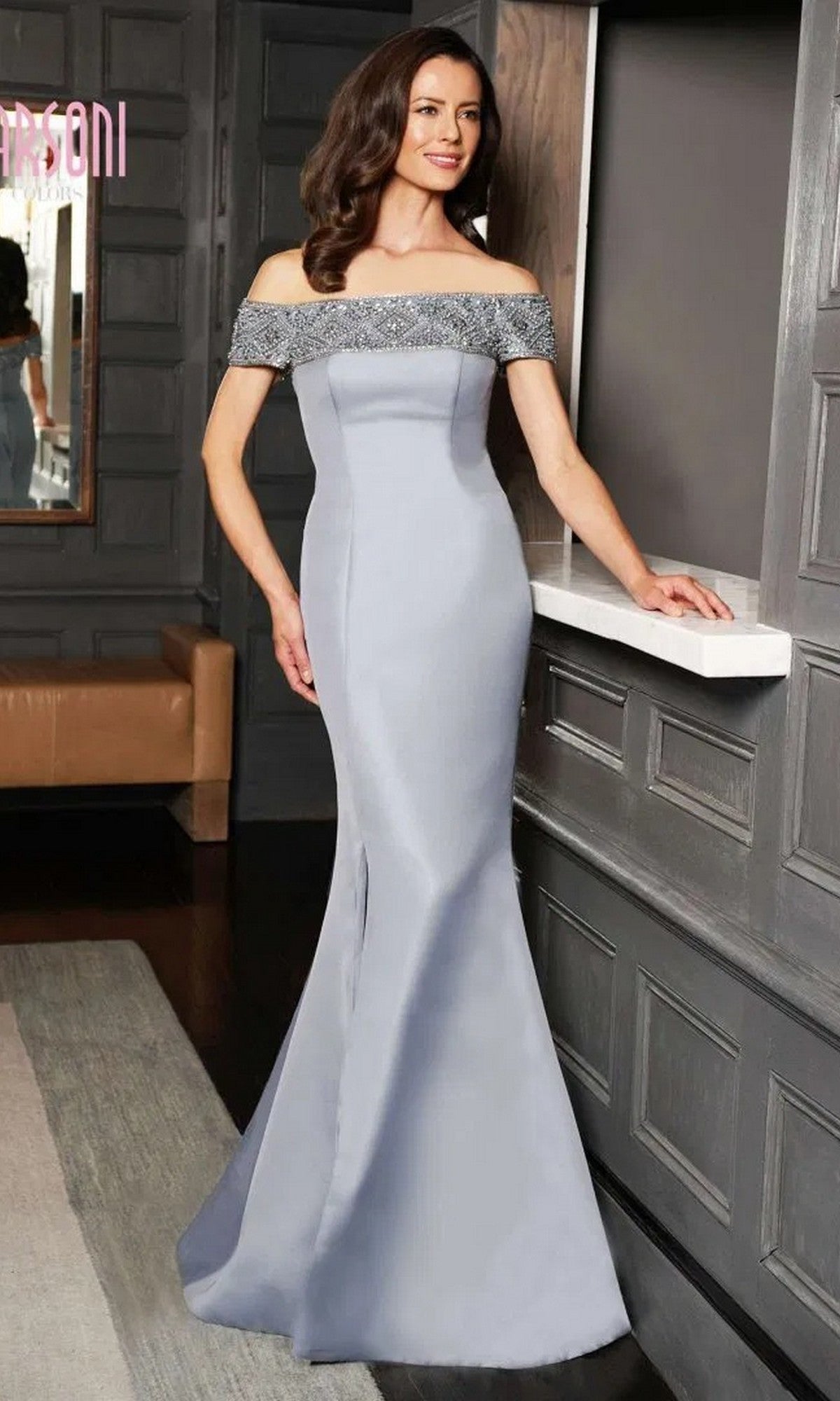 A woman in a fitted, off-shoulder Marsoni MV1184 formal floor-length dress in Wedgewood with a beaded neckline and mermaid skirt stands indoors, resting one hand on a counter.